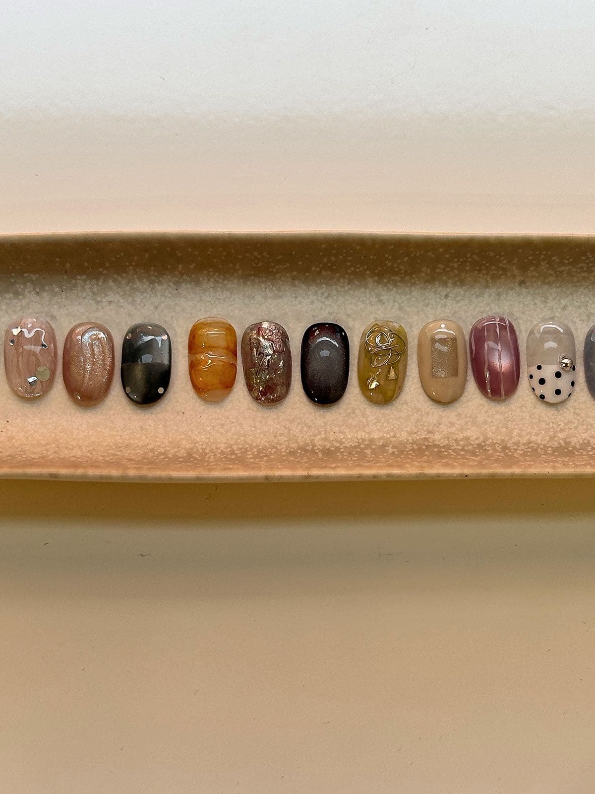 Row of earthy-toned nail art sample tips in mauve, amber and charcoal with foil and wire accents at Joy Nail Studio Vancouver