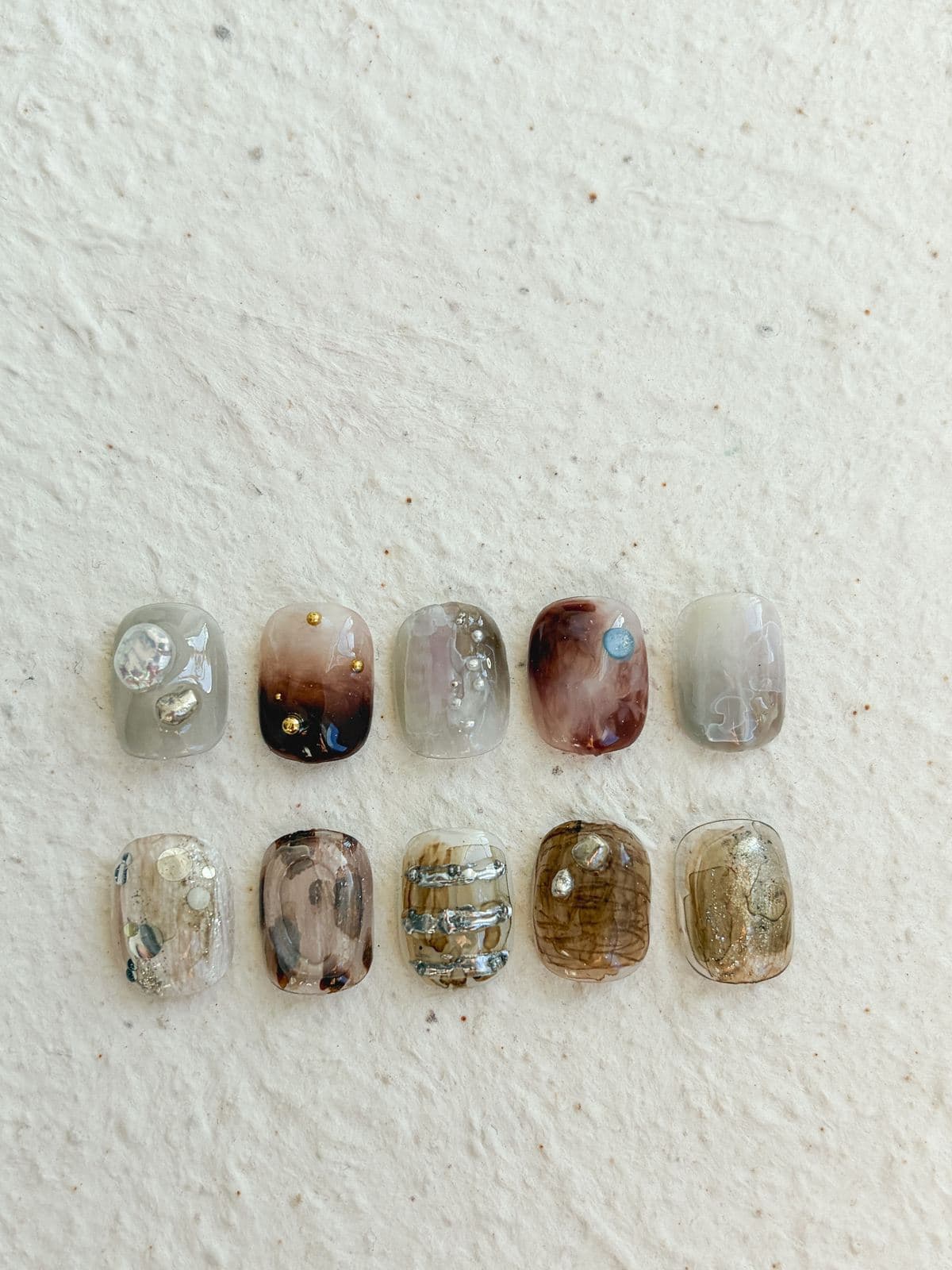 Ten October nail art sample tips in two rows on textured cream paper, tortoiseshell and brown marble with gold studs, clear tips with silver stones, raised silver bars and gold glitter, and one small blue stone, by Joy Nail Studio Vancouver