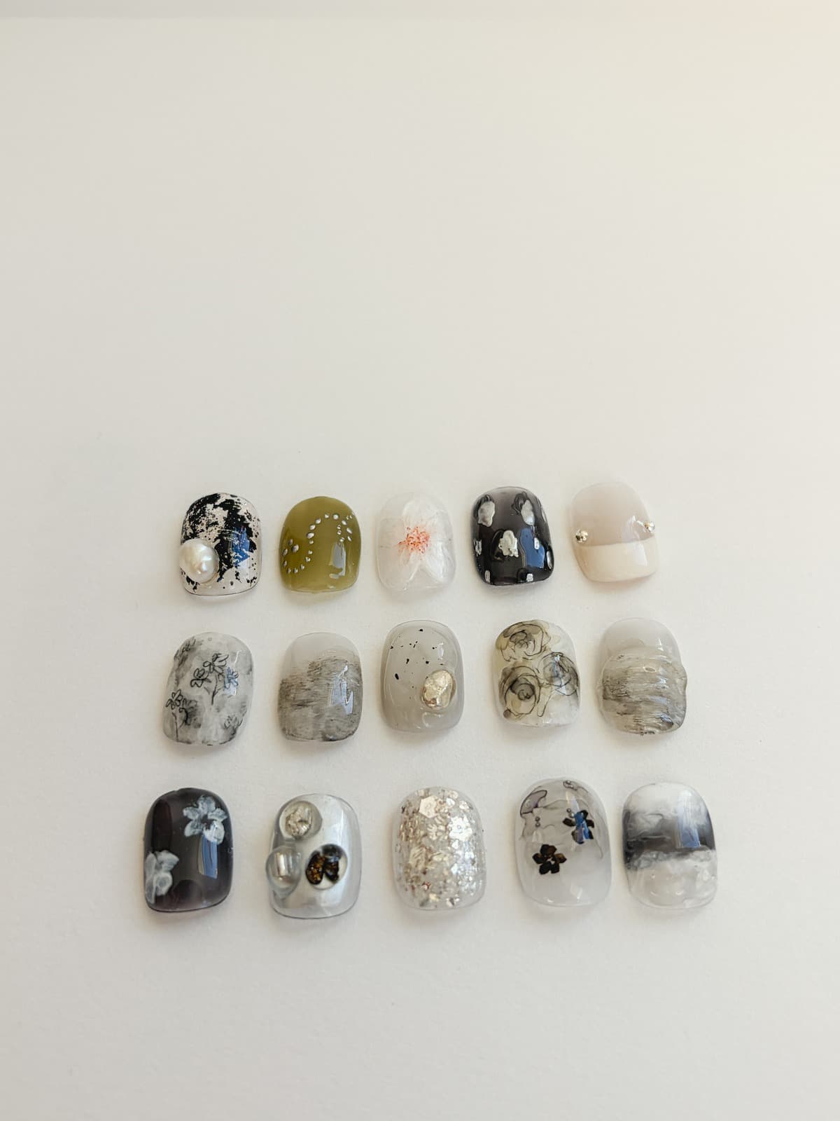 Fifteen October nail art sample tips in three rows on soft white, black-and-white marble with a pearl, olive with silver dots, grey marble with speckles, pale flowers on charcoal and black flowers on clear, silver flake and nude with silver studs, by Joy Nail Studio Vancouver