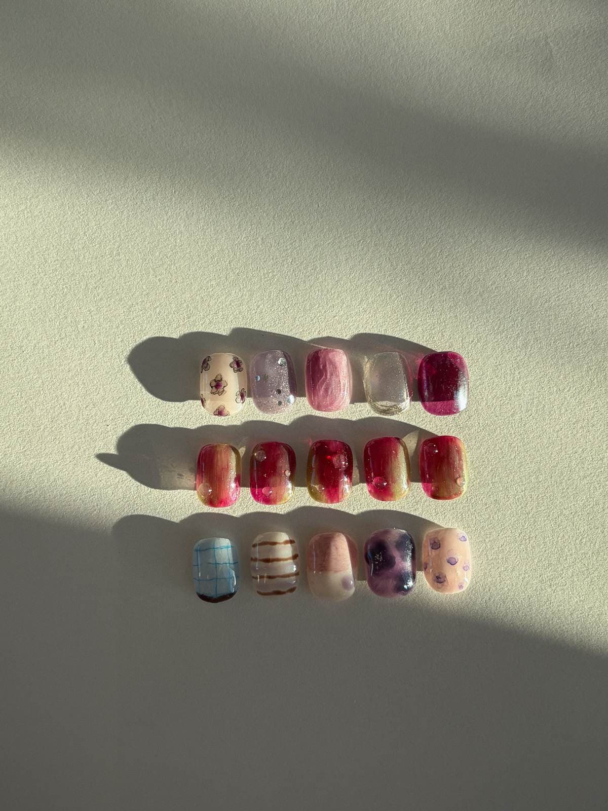The burgundy and pink nail art tips in three rows under late sun, a long shadow running across the cream plaster behind them, by Joy Nail Studio Vancouver