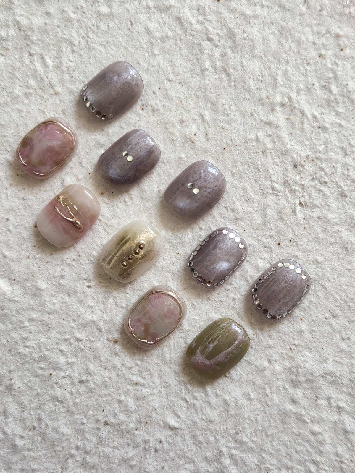 September nail art sample tips laid out loosely — pink marble, greige, pearl studs, gold foil and an olive tip on white plaster by Joy Nail Studio Vancouver